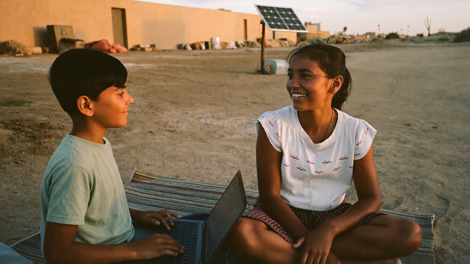 At dusk in a desert settlement, a boy works on a laptop on a woven mat while his sister talks with him; a small solar panel stands in the sand behind them.