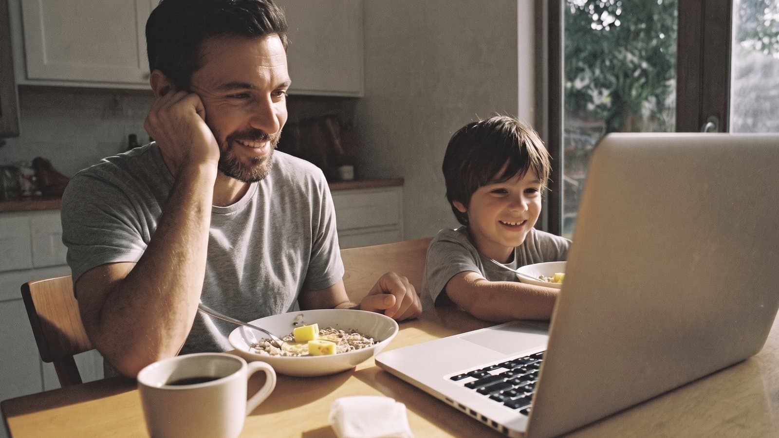 At the breakfast table, a boy works through a lesson on a laptop while his father leans on one hand beside him and smiles.