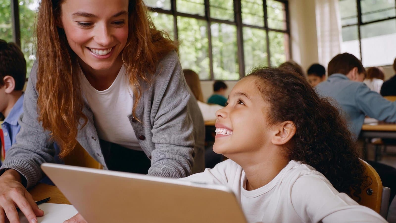 In a sunny classroom, a girl looks up from her laptop at her teacher and they share a smile; tall windows with green trees fill the wall behind them.