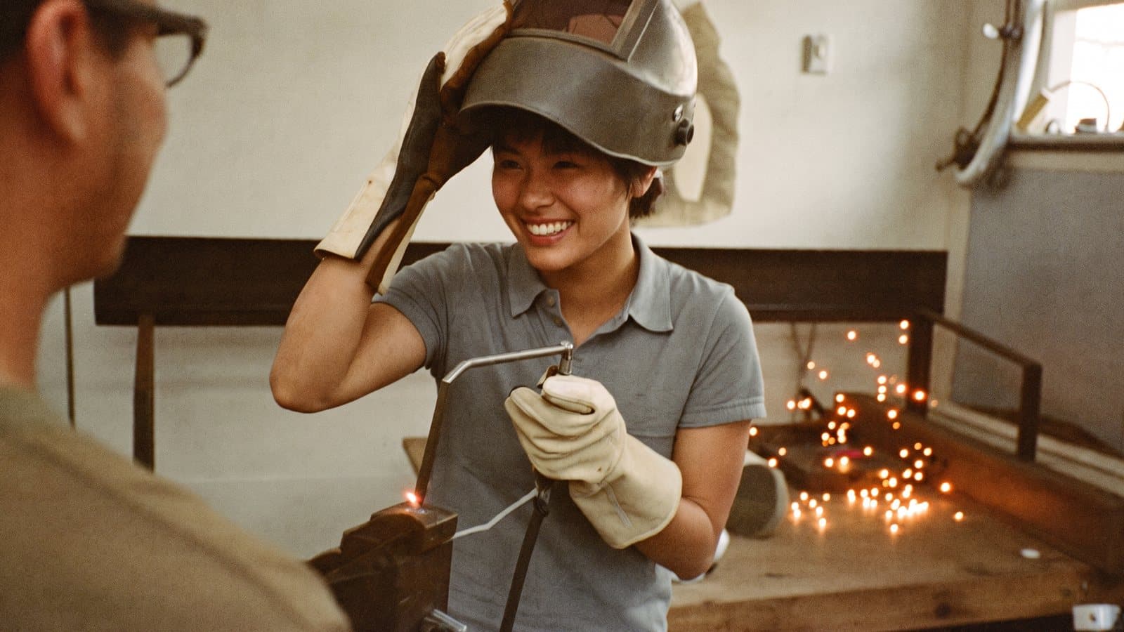 In a welding shop, a welder pushes her helmet up and grins at a coworker, sparks still glowing on the bench behind her.