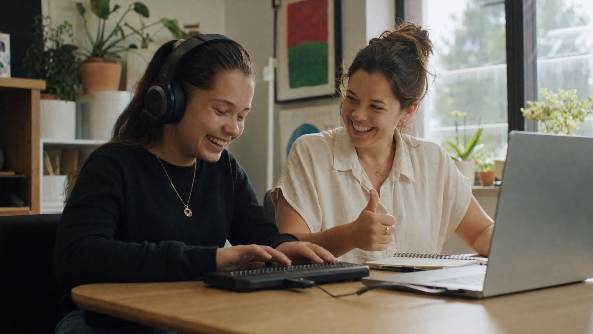 A blind teenage girl reads a lesson on a braille display and laughs as it all works, while the accessibility reviewer beside her gives a thumbs up.