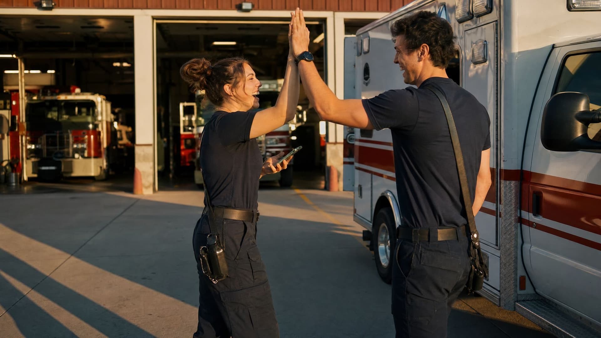 Two EMTs high-five beside their ambulance outside the fire station in the late sun.